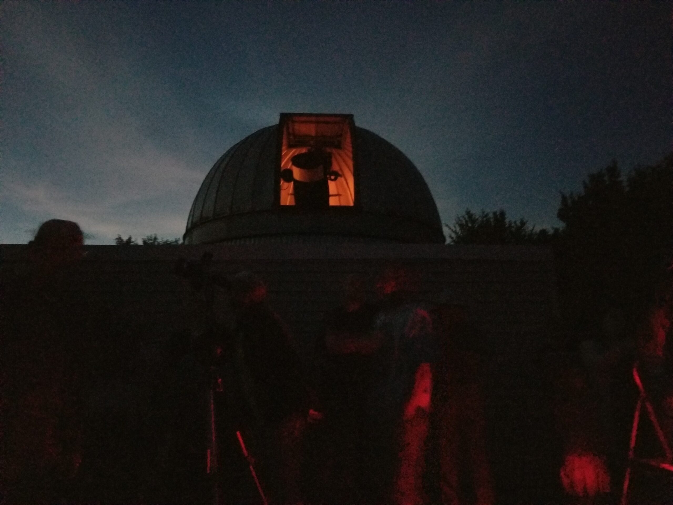 Dark image of the outside of the observatory. Red light silhouettes the telescope in the open roof.