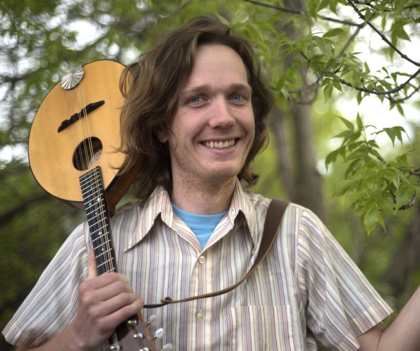 A man holding a musical instrument over his shoulder smiling outside by a tree.