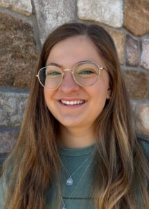 Woman with glasses smiling against a brick background.