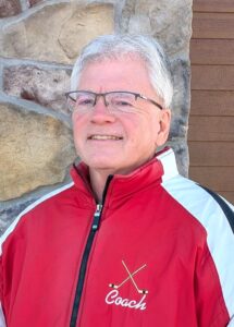 Man in a red coat, glasses and grey hair smiling against a brick background.