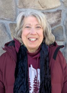 A smiling woman with grey short hair wearing a maroon jacket with a black scarf against a brick background.