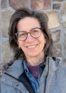 Woman smiling with short brown hair and glasses wearing a grey coat against a brick background.