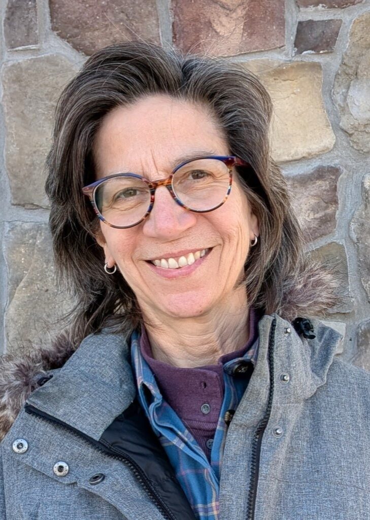 Woman smiling with short brown hair and glasses wearing a grey coat against a brick background. 