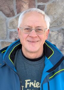 Smiling man with grey hair and glasses wearing a blue coat against a brick wall. 