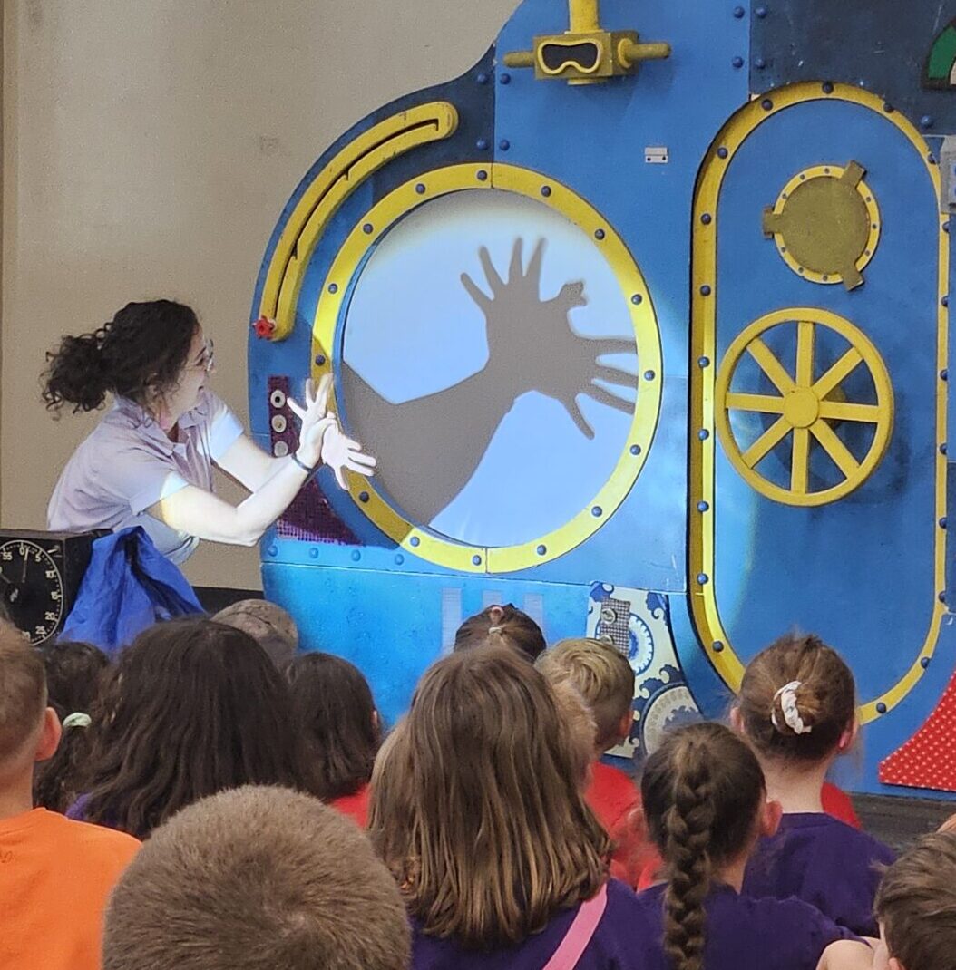 A woman standing in front of a cut out of a submarine making hand shadows on the backdrop. 