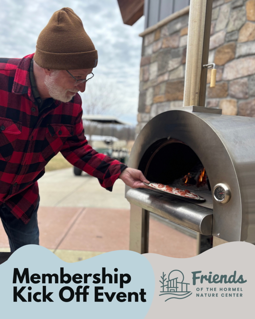 A man in a plaid jacket wearing a hat putting a pizza into an outdoor oven. 